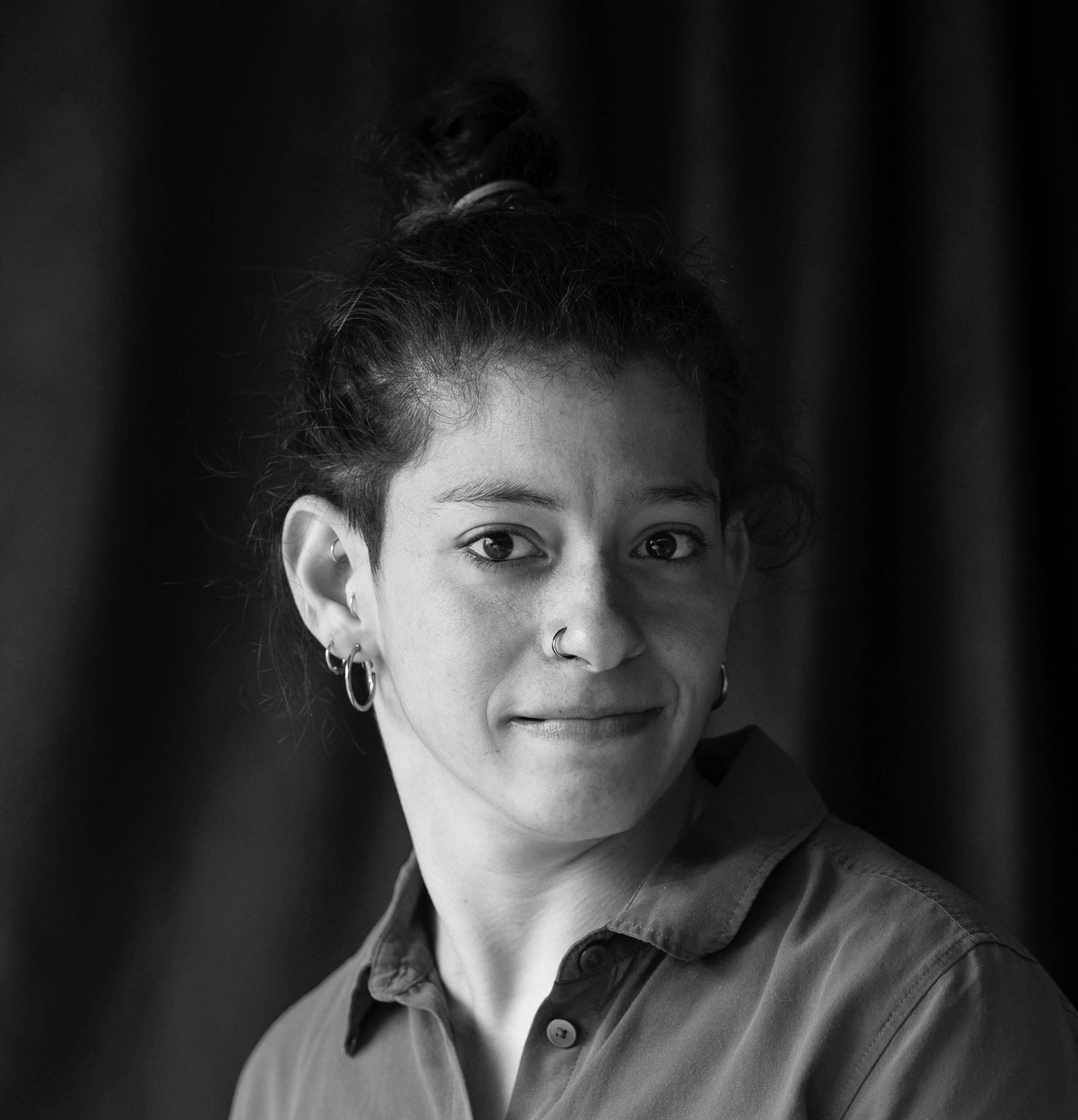 The image is a black and white headshot of Georgia Cranko. Georgia has dark curly hair tied up in a bun. They have a nose ring and are wearing hoop earrings. They are looking directly at the camera with a slight smile and are dressed in a collared shirt. The back ground is dark and out of focus making the person stand out more prominently.