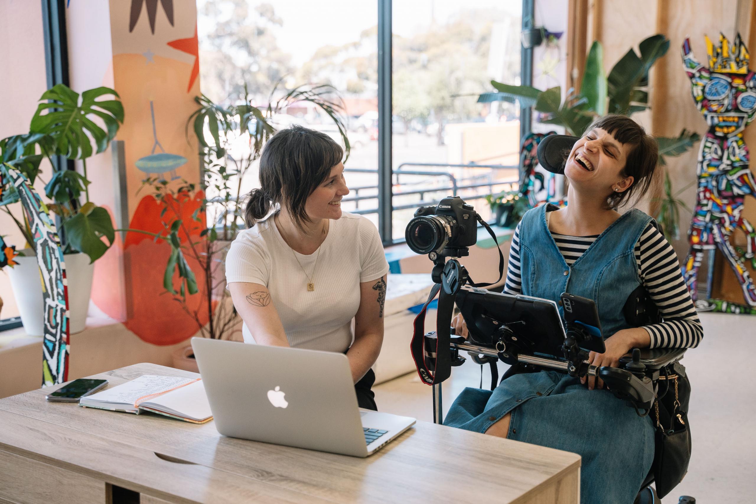 Hannah, a white woman with brown hair, sits next to Nicole, who is a woman in a wheelchair with a camera attached to it. Nic has olive skin and brown hair and is wearing a denim dress over a long sleeved striped top. Hannah is wearing black pants and a white t-shirt.