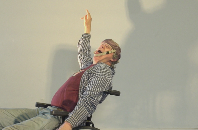 The image is a photograph of a person sitting in a wheelchair, leaning back, and pointing upwards with their right hand. The person is wearing a headset, a checkered shirt over a maroon t-shirt, and light colored pants.