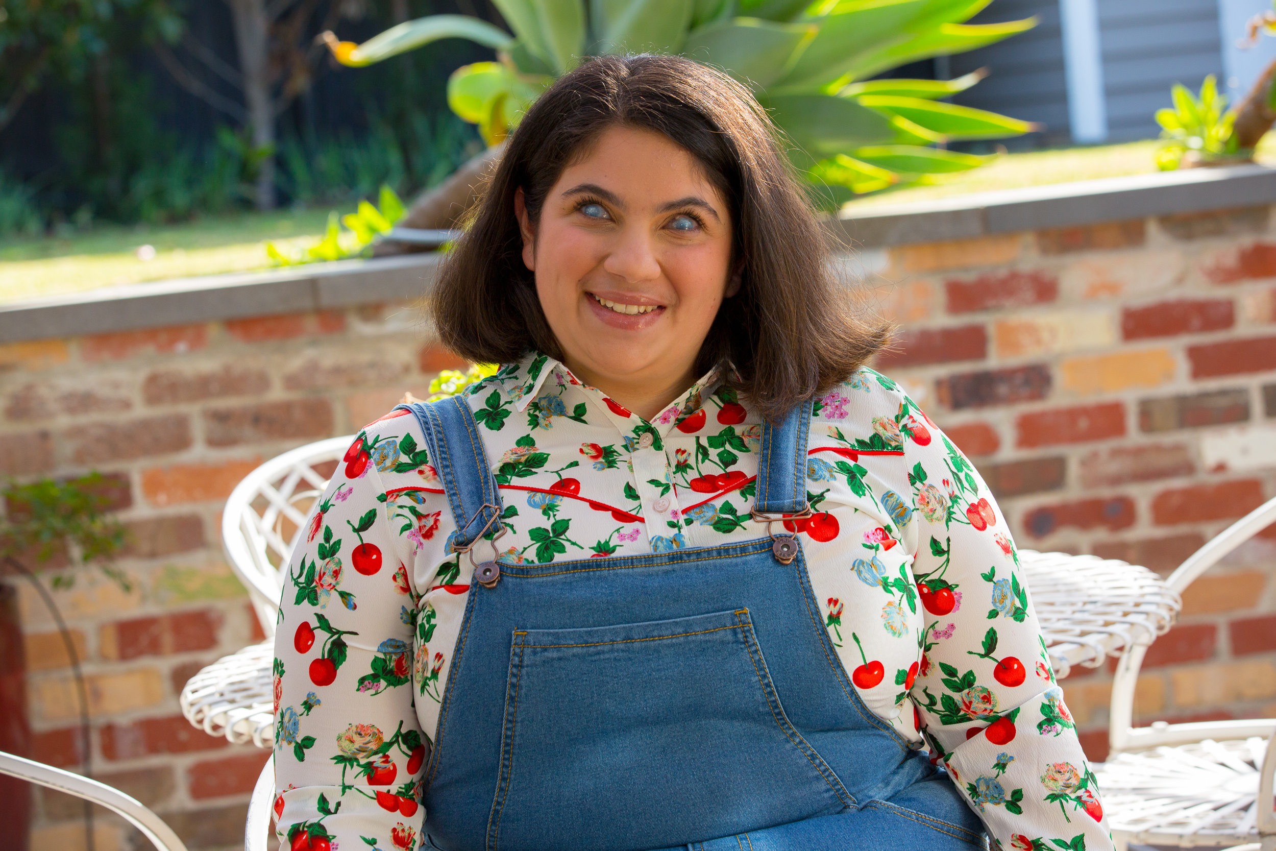 A headshot photo of Olivia who is smiling and is wearing her dark brown shoulder length hair out and parted to one side. She is wearing a white button-up shirt with a print of red cherries, green leaves and soft pink and soft blue flowers all over it. Behind her is a white outdoor furniture set and a low red brick wall with a green fern above it. Blurred into the background behind the fern is a garden with green grass.