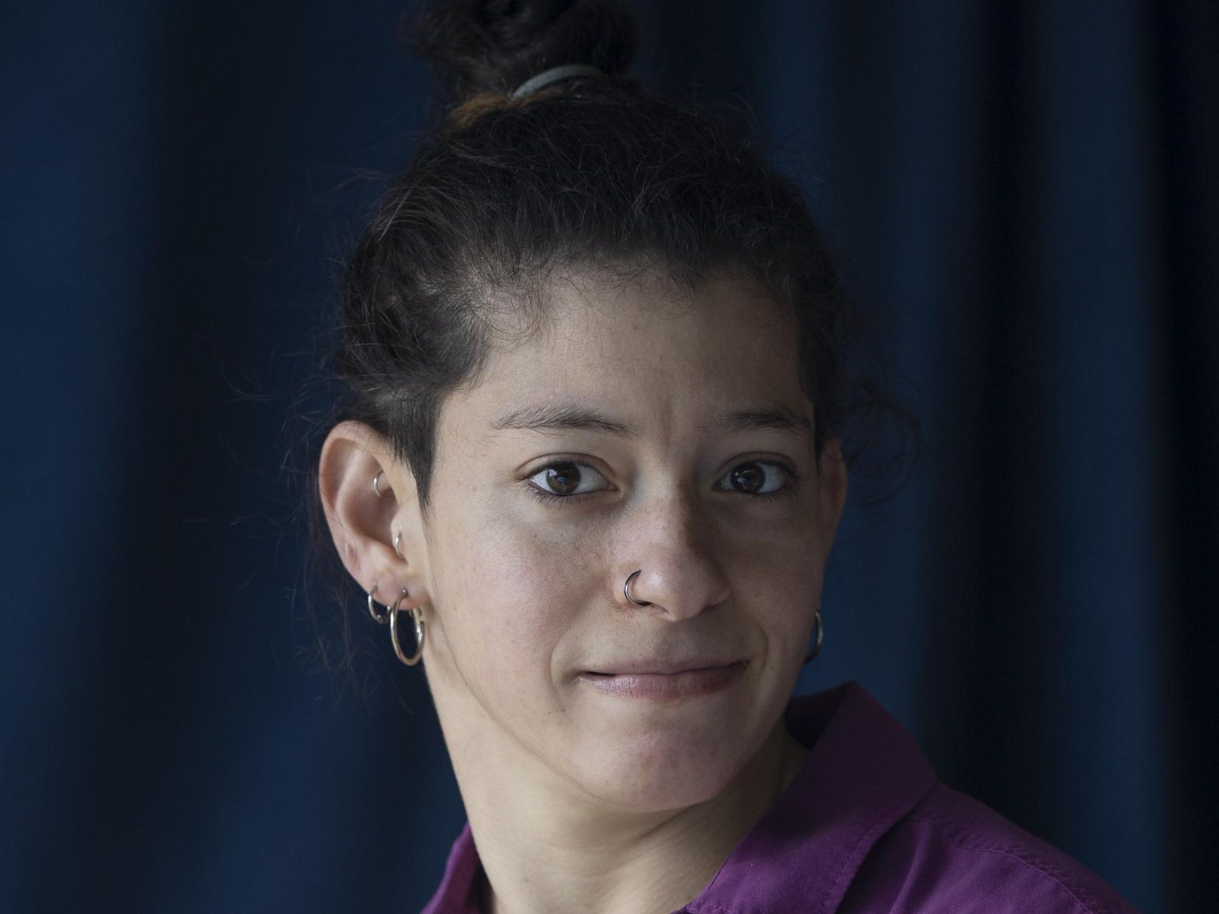 The image is a headshot photo of Georgia Cranko. They have dark hair that is tied up in a bun. They have a light complexion and are wearing a purple collared shirt. They have a nose ring and small hoop earrings in both ears. Georgia is looking slightly to the side with a neutral expression on their face. The background is dark blue.