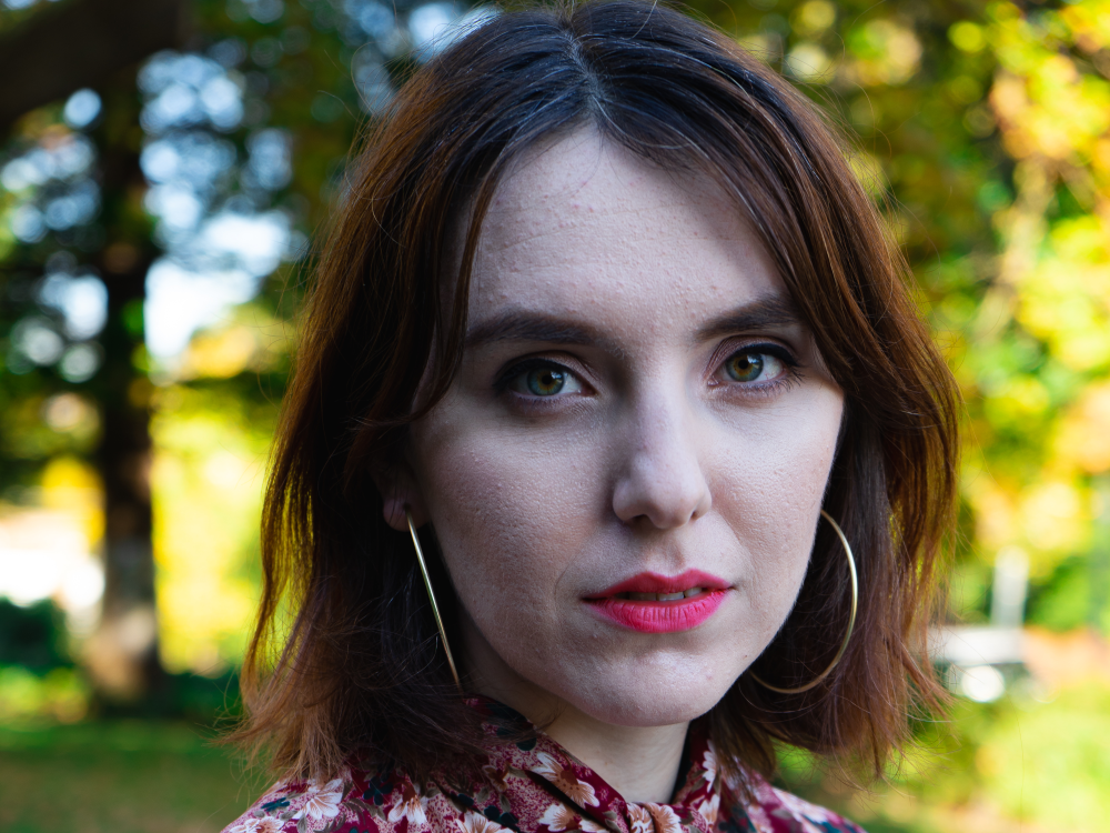 The picture is a headshot photo of Tash Atkins who has fair skin and shoulder-length brown hair. They are wearing a floral blouse with a red background and flowers in shades of pink and white. They are wearing large hoop earrings and are looking directly at the camera with a neutral expression. The background is an outdoor setting with green trees and a pathway on a sunny day.