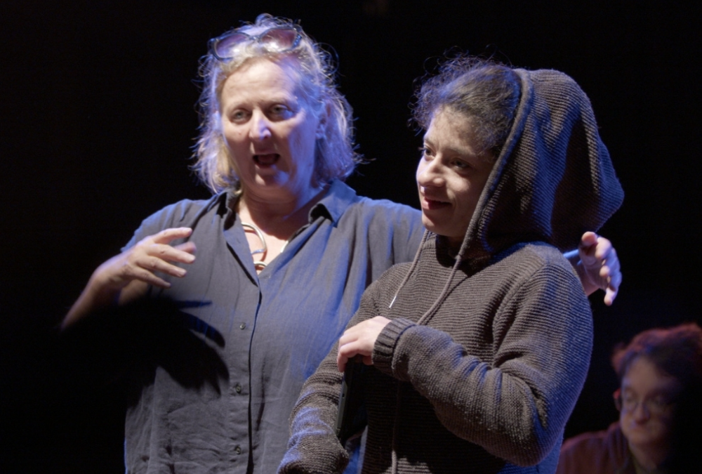 The image is a photo of director Jenny Sealey and Georgia Cranko standing close together, engaged in conversation. Jenny is on the left and is an older woman with light grey shoulder length hair, wearing glasses on her head and is dressed in a dark loose fitting charcoal shirt. She has an expressive face and her mouth is open as if she is speaking. Georgia is on the right with darker hair, pulled back and is wearing a hoodie. They are smiling and looking towards Jenny. In the background, there is another person, Matt, who is slightly out of focus wearing glasses and also dressed in dark clothing. The setting appears to be a stage.