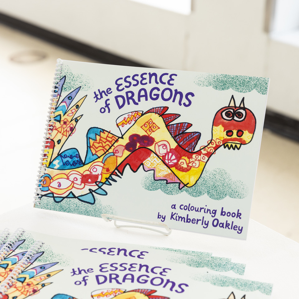 A photograph of Kimmy’s colouring book on a white stand. The cover features a multi-coloured hand-drawn dragon with a red head, big eyes, horns, and many curved spikes. The background is mint with teal clouds. The text on the cover is purple and reads ‘The Essence of Dragons, a colouring book by Kimberly Oakley’. The book is bound with a white plasticoil spiral.