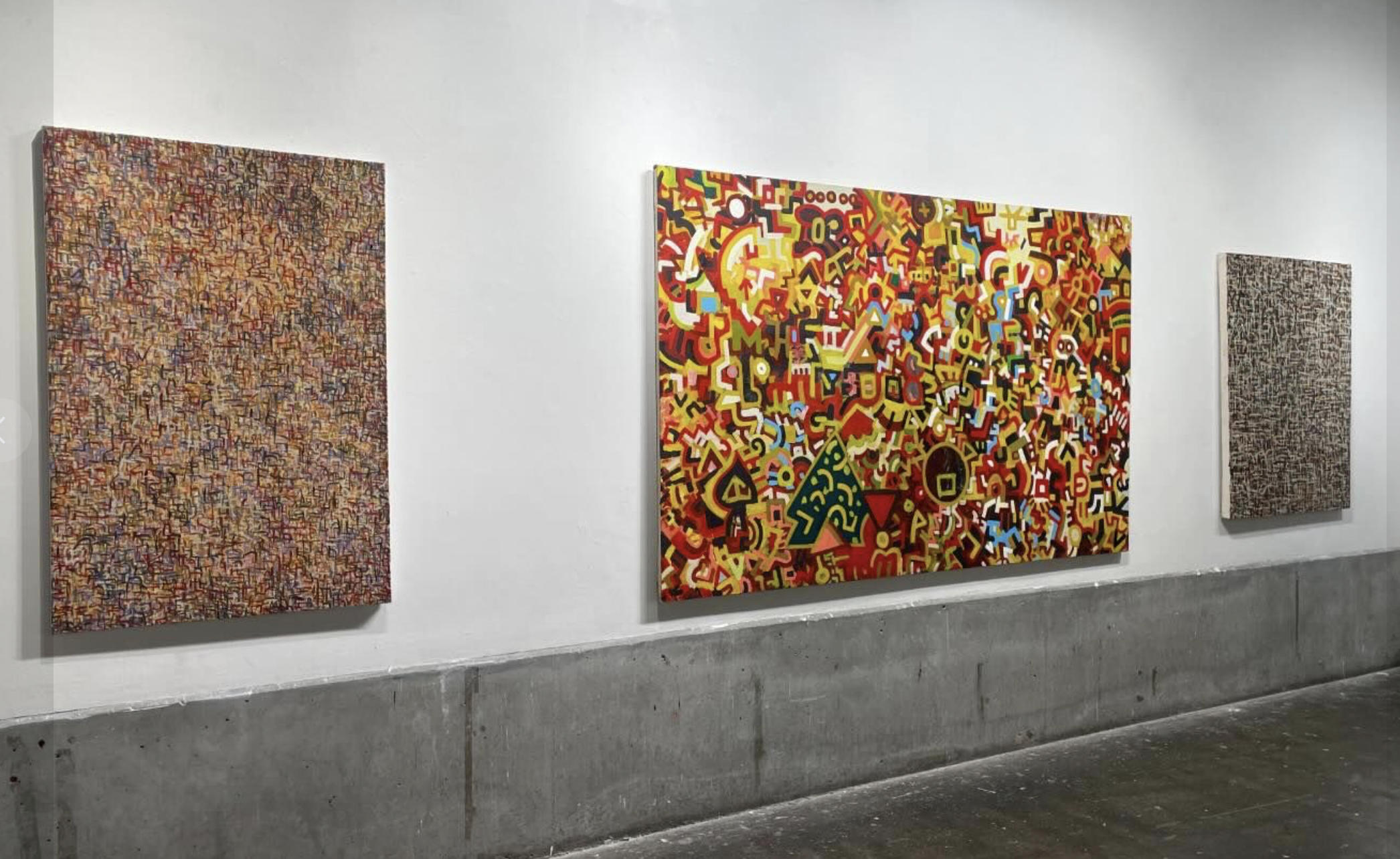 The image is a photo of an art gallery wall. The wall displays three abstract paintings in a row. The painting to the left is a densely packed mosaic of small, colorful shapes and lines creating a textured, intricate pattern. The middle painting is larger and features a vibrant chaotic mix of bright colors and abstract shapes, including reds, yellows and blues. The painting to the right is similar to the one on the left, with a dense arrangement of small, colorful shapes and lines. All paintings are by Matthew Simpson.
