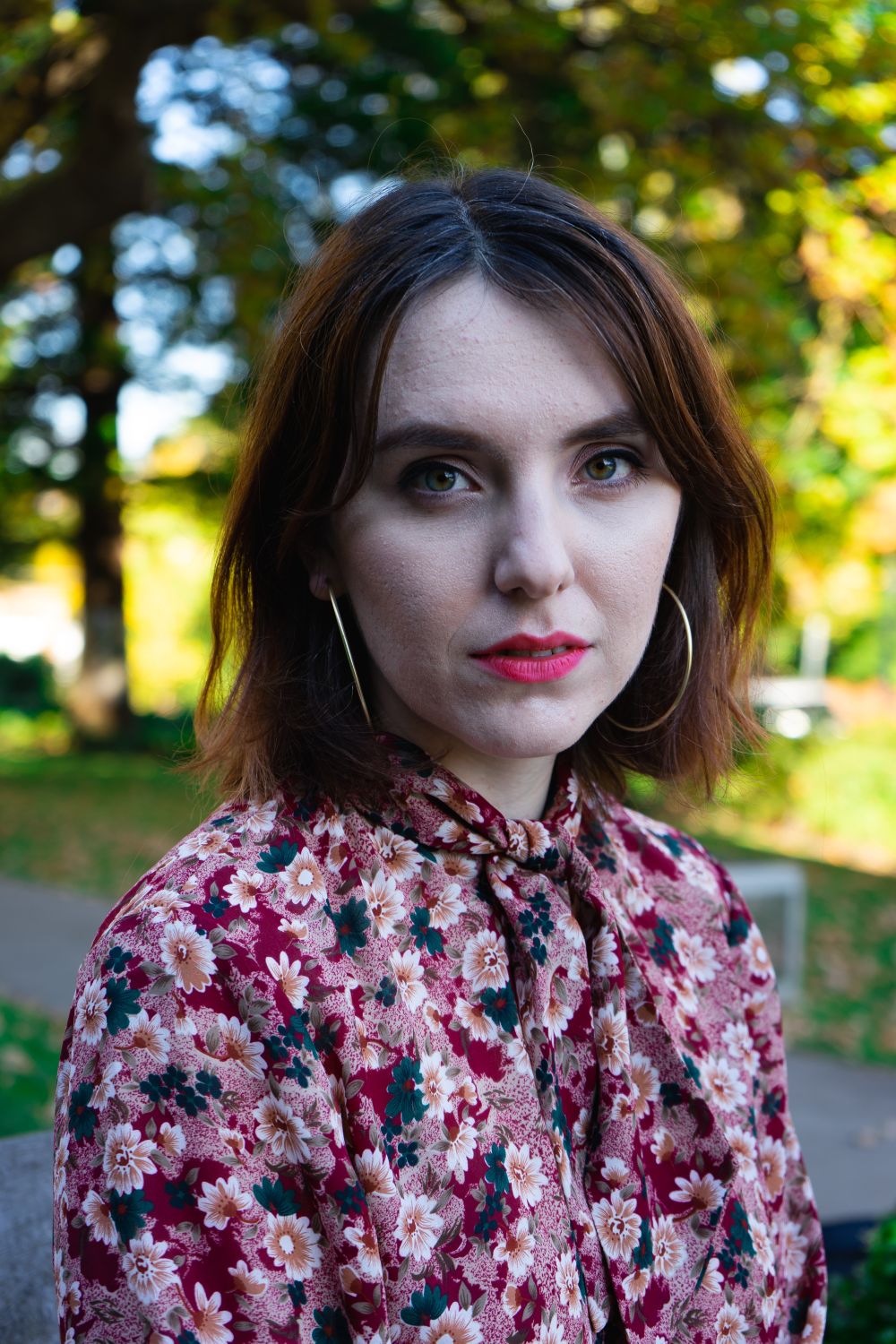 The picture is a headshot photo of Tash Atkins who has fair skin and shoulder-length brown hair. They are wearing a floral blouse with a red background and flowers in shades of pink and white. They are wearing large hoop earrings and are looking directly at the camera with a neutral expression. The background is an outdoor setting with green trees and a pathway on a sunny day.