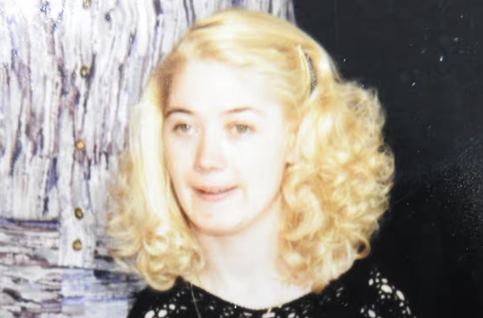 The image is a photo of Ann Marie Smith, a young woman with fair skin and light blonde, curly shoulder length hair. She is wearing a black top with a lace-like pattern and has a slight smile on her face. The background appears to be a patterned fabric with vertical stripes in various shades of grey and white.