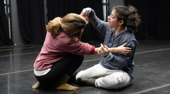 The image shows two people engaged in a dance or movement exercise. They are both on the floor, facing each other. The woman on the left (Amanada McGregor) is wearing a pink sweater and has her hair tied back. She is crouched down with her arms extended towards the other person. The person on the right (Georgia Cranko) is wearing a blue hoodie and great track pants. They are sitting on the floor with their legs bent and are also extending their arms towards Amanda. Amanda and Georgia are holding each other's hands and seem to be in a moment of connection or interaction. The background is a dark indoor dance studio with black curtains.