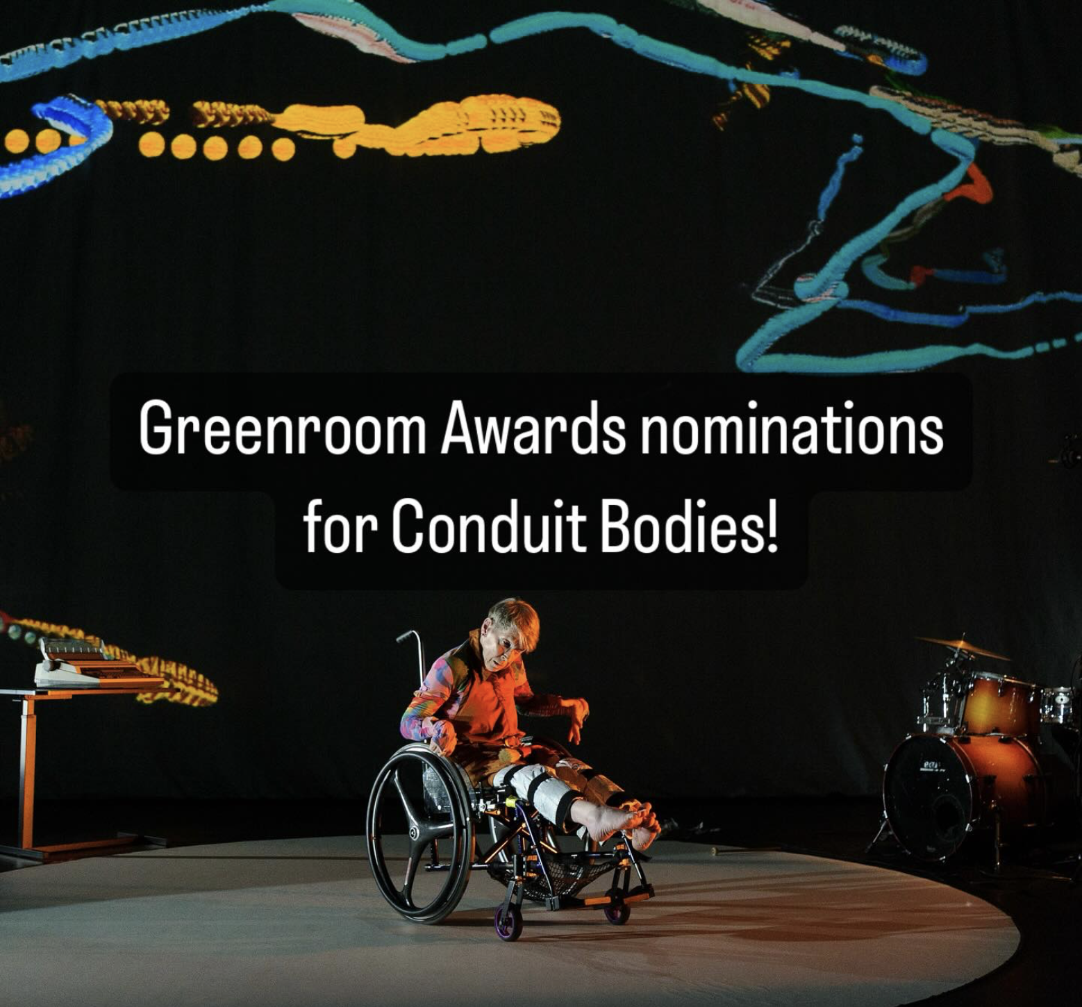A performance shot of Mel Smith in a wheelchair on a circular stage, wearing terracotta coloured overalls. Abstract patterns of squiggly lines and circles are projected on a dark background in blue and yellow. A table with typewriter is on the left, and a drum set is on the right. Dramatic lighting highlights the scene. White text in the centre reads: Greenroom Award nominations for Conduit Bodies!
