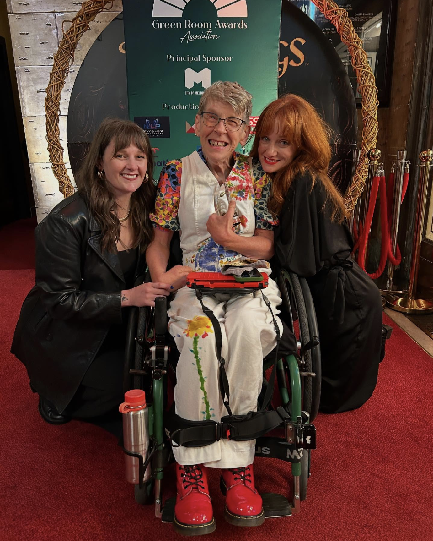 A photo of Melinda Smith holding an award and smiling, with Hannah Reekie and Zoe Boesen either side of her. Mel wears white overalls with a hand painted yellow flower on her right knee. On the left, Hannah who has long dark hair and in a black leather jacket smiles, and on the right, Zoe wearing a black jumpsuit also smiles. Behind them is a “Green Room Awards Association” banner.