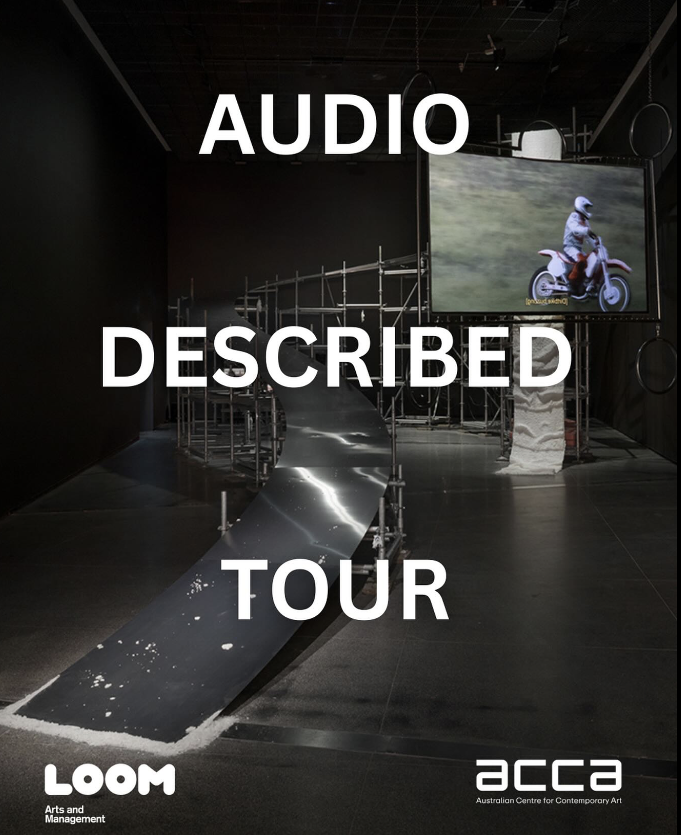 a dark exhibition space with a large art installation and white text overlaid. A large silver ramp supported by metal scaffolding curves around the back of the room, from floor to almost ceiling height on the right. In front of the ramp to the right, a large rectangular screen is suspended from the ceiling. It shows a person riding a white motorcycle outdoors, with white protective clothing and helmet. Overlaid text reads: AUDIO DESCRIBED TOUR. Loom and ACCA logos are at the bottom.