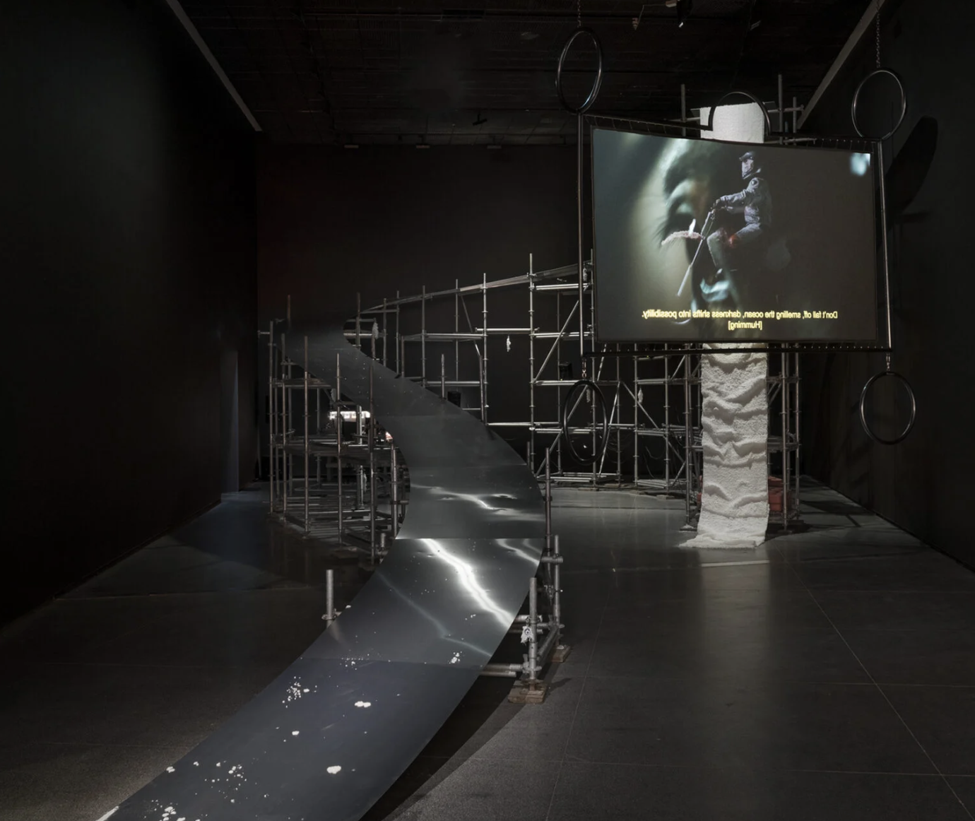 a dark exhibition space with a large art installation and white text overlaid. A large silver ramp supported by metal scaffolding curves around the back of the room, from floor to almost ceiling height on the right. In front of the ramp to the right, a large rectangular screen is suspended from the ceiling. It shows a person riding a white motorcycle outdoors, with white protective clothing and helmet.