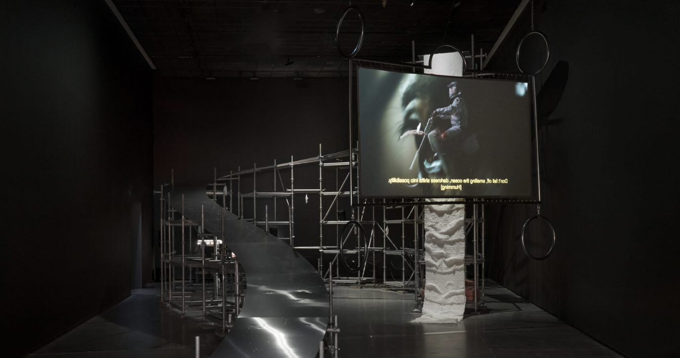 a dark exhibition space with a large art installation and white text overlaid. A large silver ramp supported by metal scaffolding curves around the back of the room, from floor to almost ceiling height on the right. In front of the ramp to the right, a large rectangular screen is suspended from the ceiling. It shows a person riding a white motorcycle outdoors, with white protective clothing and helmet.