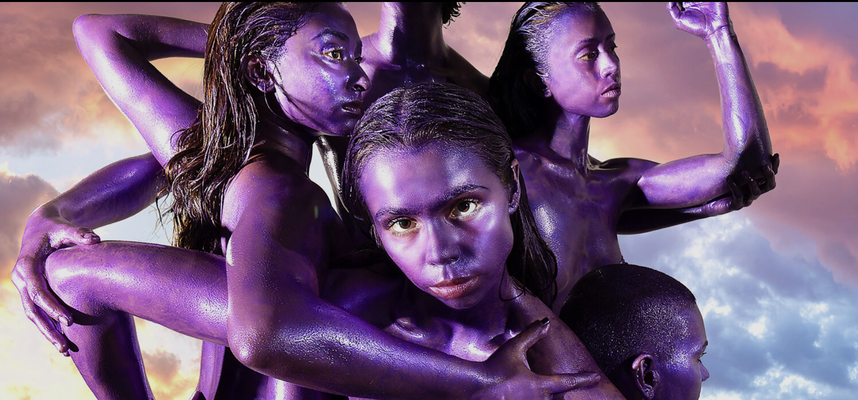A still image of 4 women with purple-painted skin, intertwined and posing closely together. Their bodies and faces are covered in shiny, metallic purple paint, and they are looking in different directions with serious expressions. The background is a dramatic sky with clouds lit by a sunset or sunrise, casting an orange and pink glow.