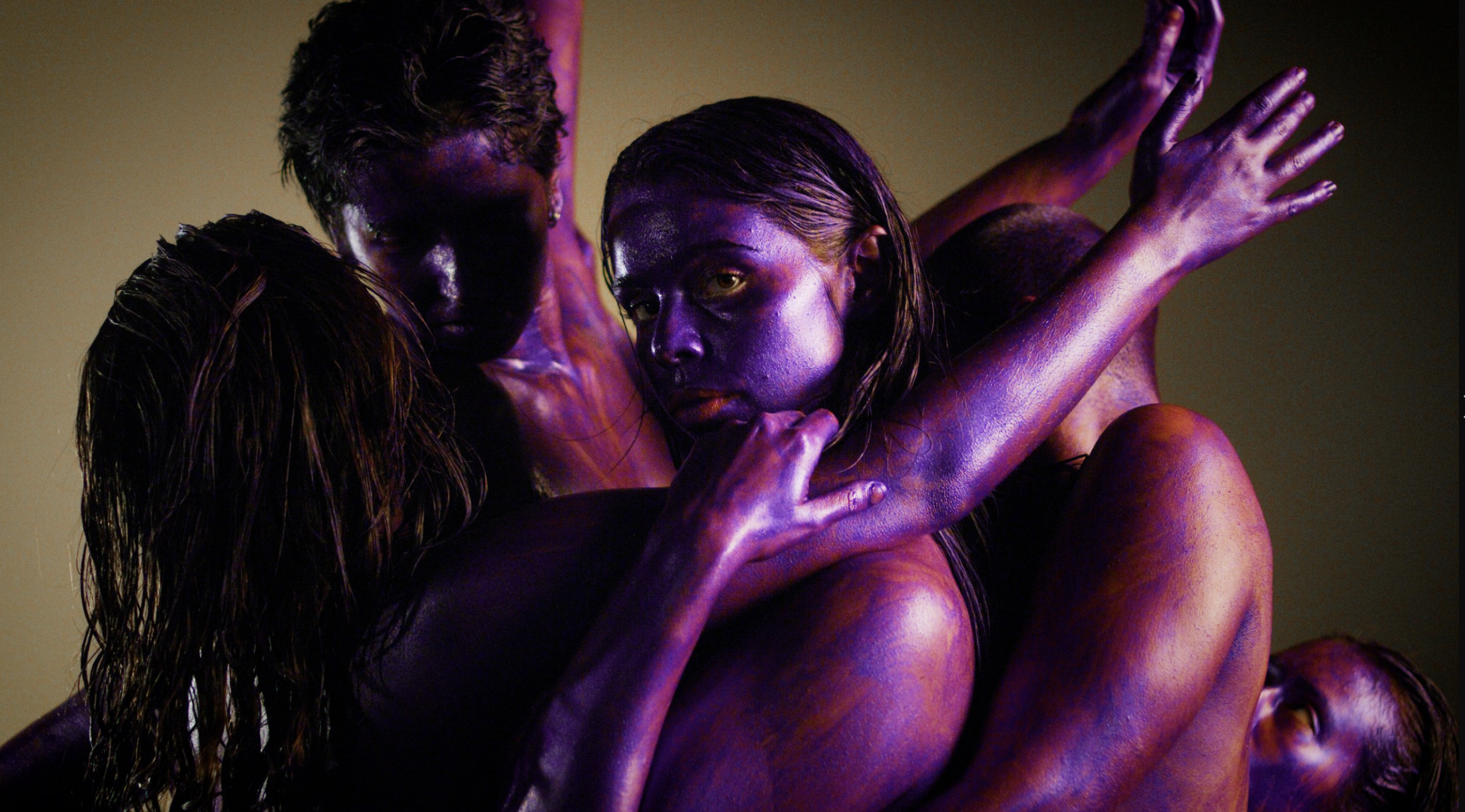 A still image of 4 women with purple-painted skin, intertwined and posing closely together. Their bodies and faces are covered in shiny, metallic purple paint, and they are looking in different directions with serious expressions. The background is a deep gold colour.
