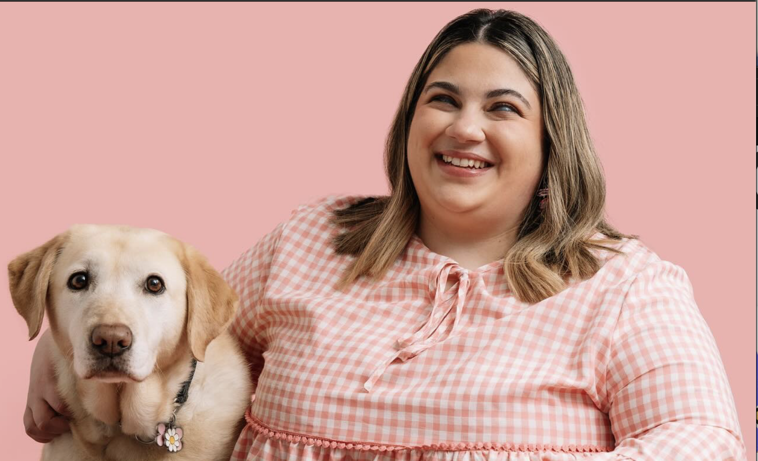Olivia, a woman with a fair complexion and dark hair with blonde highlights, and Jemima, a yellow Labrador sitting together at a pink table. Olivia is wearing a pink and white gingham-patterned dress and is smiling widely. Her right arm is around Jemima, who is sitting calmly and looking directly at the camera. The background and table are both solid pastel pink.