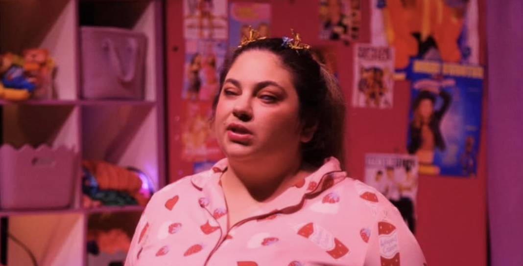 Olivia stands, dressed in pink pajamas with red heart and cupcake designs, in a colorful bedroom illuminated with pinkish ambient light. She is facing forward and appears to be mid- speech or mid-song. Her hair is tied back into a ponytail and she has colourful butterfly clips in her hair. The background features cube shelves filled with toys, clothing and storage bins, with posters and magazine cutouts decorating the walls. The overall scene feels playful and cozy, depicting a youthful and whimsical atmosphere.