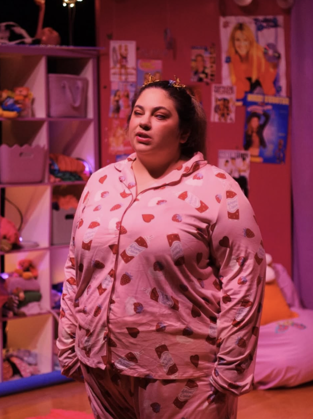 Olivia stands, dressed in pink pajamas with red heart and cupcake designs, in a colorful bedroom illuminated with pinkish ambient light. She is facing forward and appears to be mid- speech or mid-song. Her hair is tied back into a ponytail and she has colourful butterfly clips in her hair. The background features cube shelves filled with toys, clothing and storage bins, with posters and magazine cutouts decorating the walls. The overall scene feels playful and cozy, depicting a youthful and whimsical atmosphere.