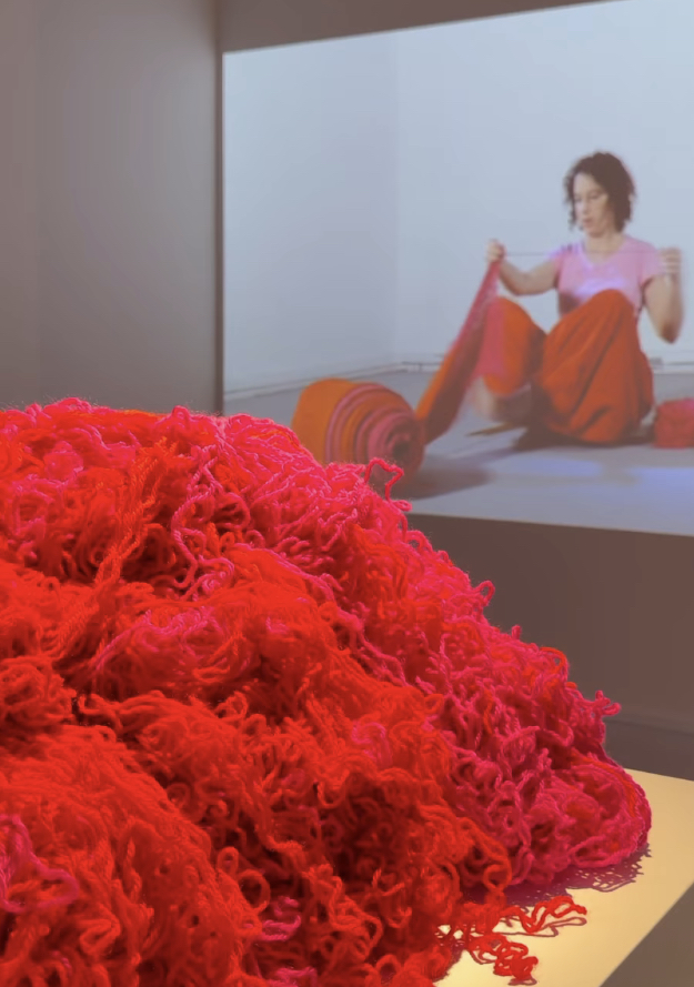 A photo of Prue Stevensons work 'By- Product'. In the foreground is a pile of red and pink yarn. In the background is a projection on the gallery wall of Prue sitting cross legged and unraveling wool from a spool.