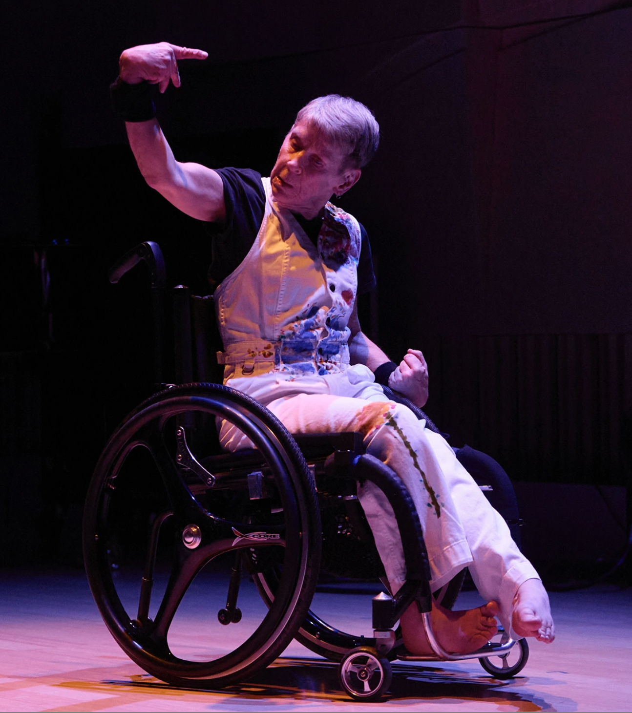  Dr Melinda Smith is in a wheelchair onstage, illuminated by dramatic lighting with pink and purple hues. The background is dark highlighting the performer. Mel is photographed mid dance move caught in a strong, expressive pose: one leg slightly extended from the chair, their left hand clenched into a fist at their waist and the other arm raised and pointing over her head. Her facial expression looks intense and focussed. Mel is wearing a black t shirt under white overall onto which she has painted designs across the shoulder, stomach and down the legs. She is also wearing artist bands which contain motion sensors which are used to control the visuals and music onstage during the performance.